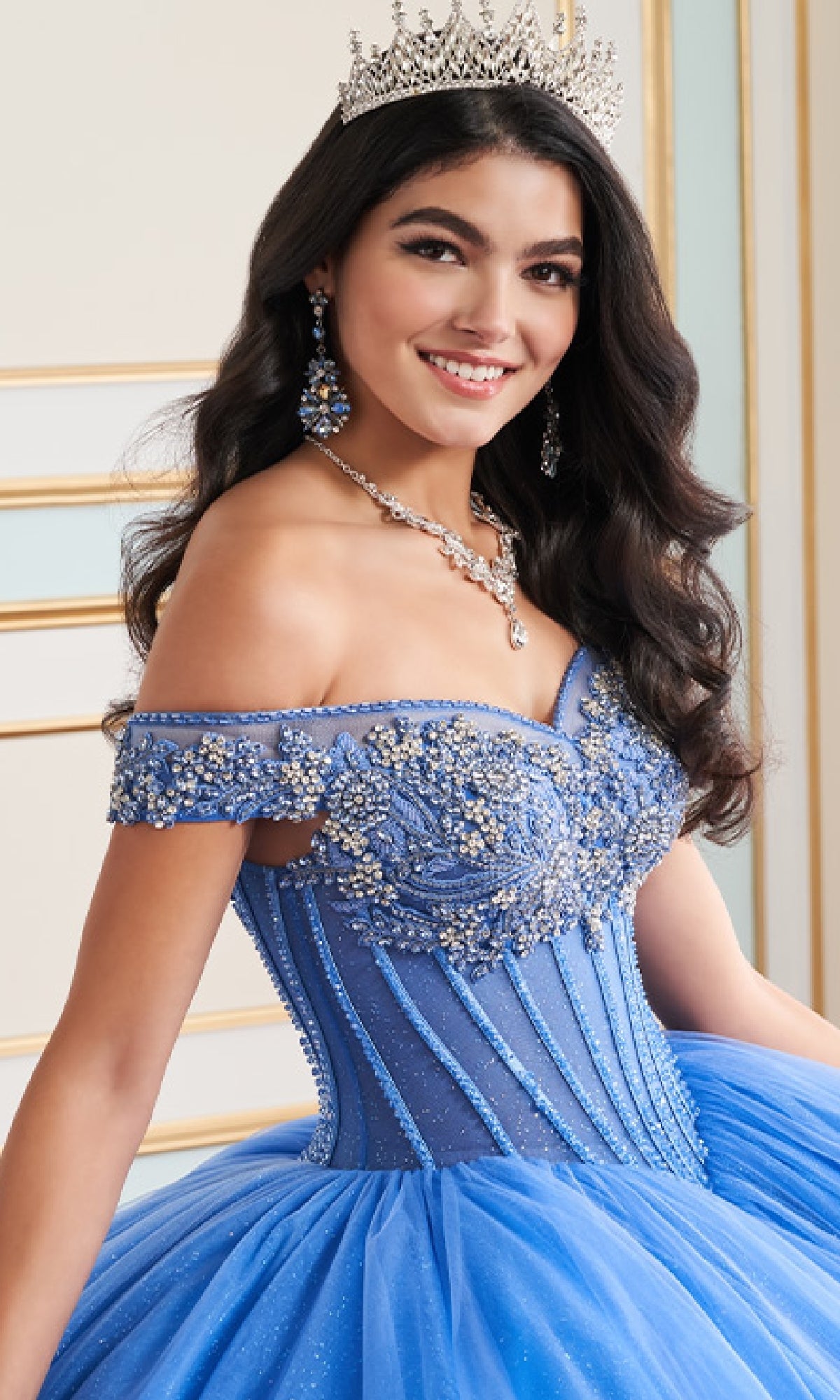 A woman stands indoors, smiling at the camera in the Quinceanera Dress PR30180 By Princesa—a jeweled blue ball gown with an off-the-shoulder neckline—styled with a silver crown and matching jewelry.